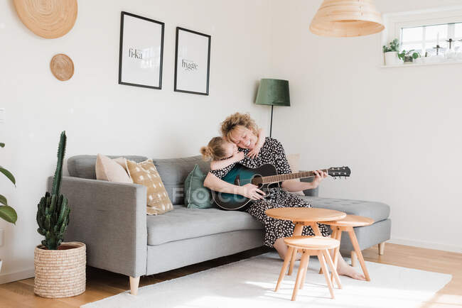 Дочка дає мамі обійми, в той час як вона грає на гітарі вдома — стокове фото