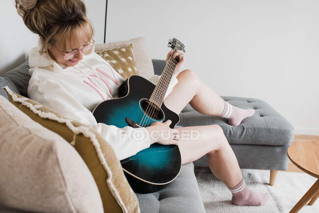 Femme assise à jouer de la guitare à la maison sur le canapé — Photo de stock