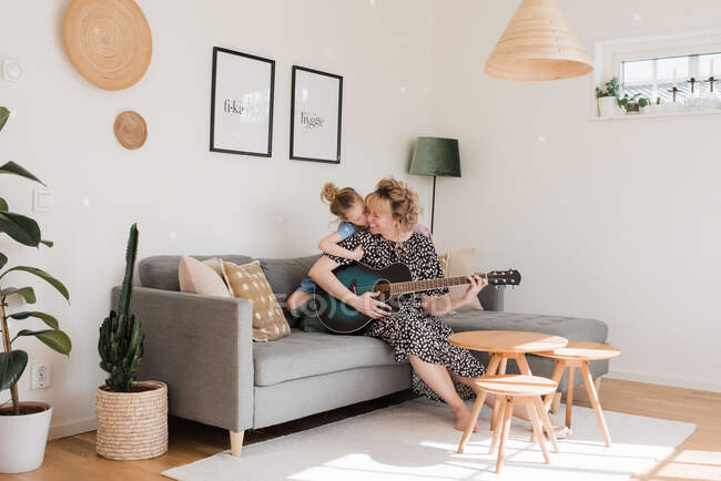 Hija besando a su mamá mientras ella toca la guitarra en casa - foto de stock