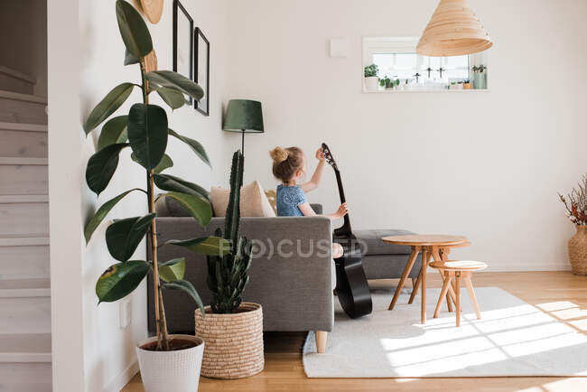 Jeune fille accordant sa guitare alors qu'elle était assise sur le canapé à la maison — Photo de stock