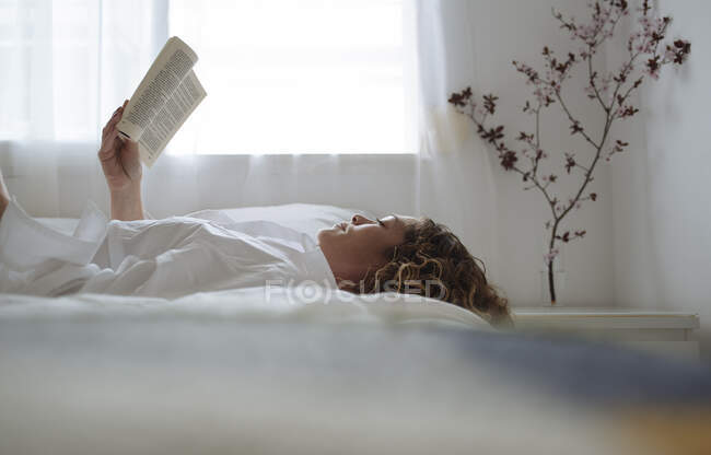 Donna dai capelli ricci che legge un libro sdraiato sul letto — Foto stock