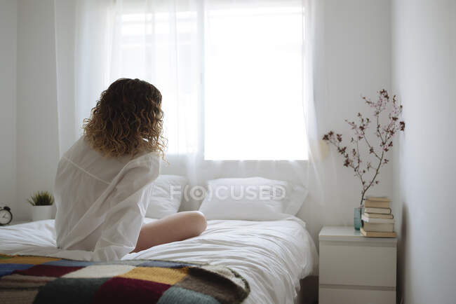 Donna in camicia bianca seduta di nuovo alla macchina fotografica sul letto — Foto stock