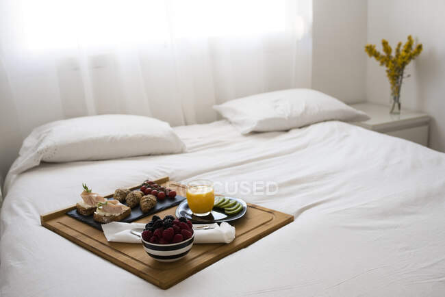 Bandeja de desayuno con fruta y tostadas en una cama en una habitación blanca - foto de stock