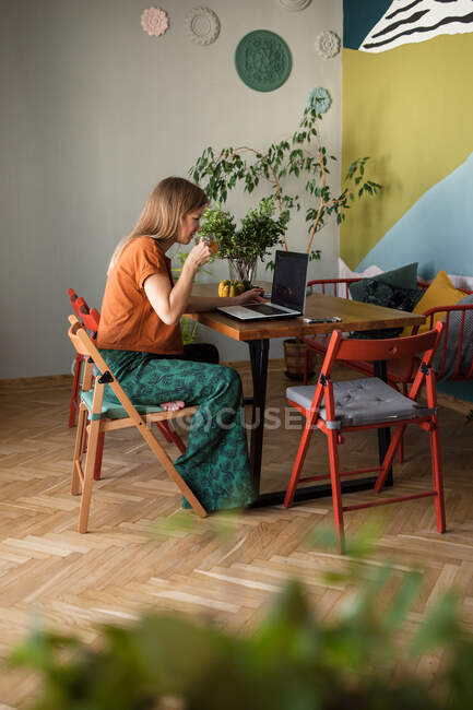 Mulher com xícara de vidro de chá funciona on-line em casa por laptop na mesa — Fotografia de Stock