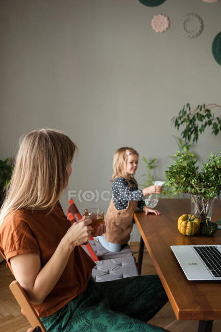 Мать с чашкой чая отдыхает и смотрит на ребенка, который поливает зеленые растения — стоковое фото