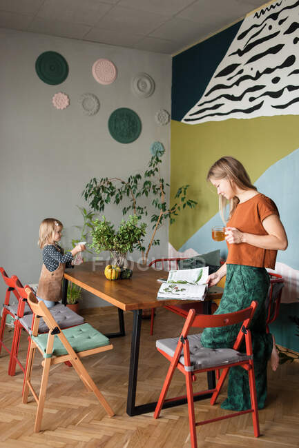 Mãe lê revista de pé à mesa, plantas de águas infantis na mesa — Fotografia de Stock