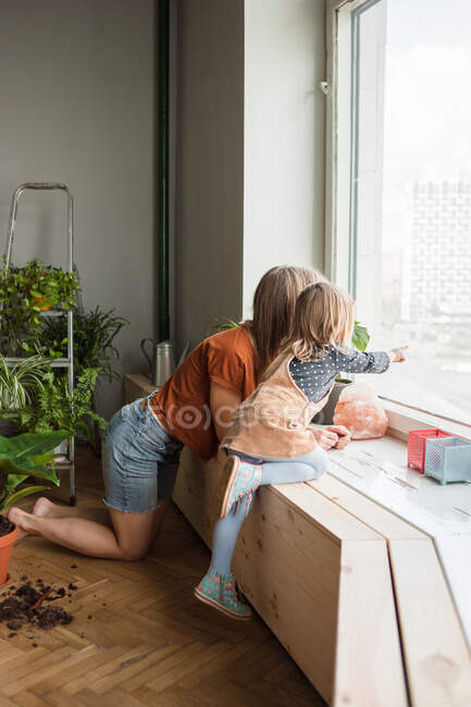 Mother and daughter look outside kneeling at window. Kid points. — Stock Photo