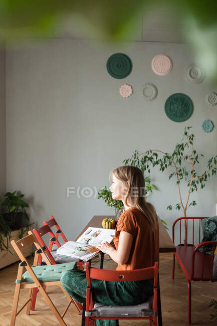 Жінка сидить за столом з журналом, дивиться на вікно у вітальні . — стокове фото