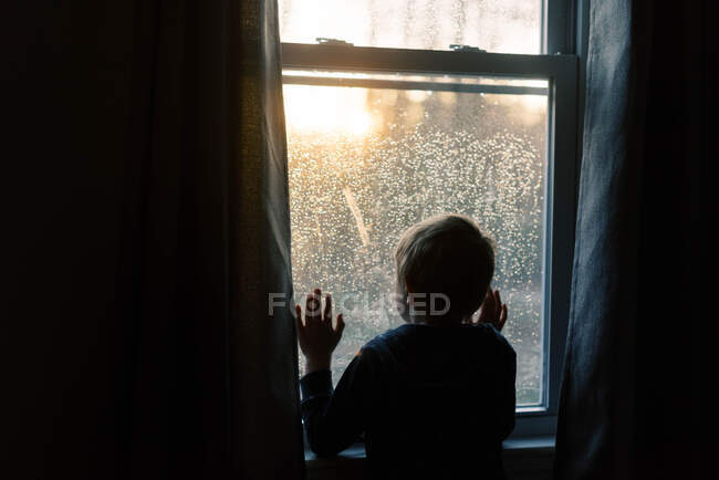 Petit garçon regardant le coucher du soleil à travers une fenêtre. — Photo de stock