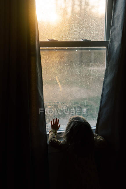 Menina criança olhando para o pôr do sol através de uma janela. — Fotografia de Stock