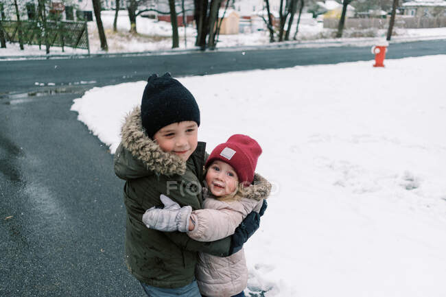 Geschwister umarmen sich in der Einfahrt beim Spielen im Schnee. — Stockfoto