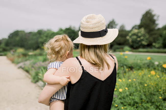 Мама в шляпе, держит малыша, пока смотрит на цветочное поле. — стоковое фото