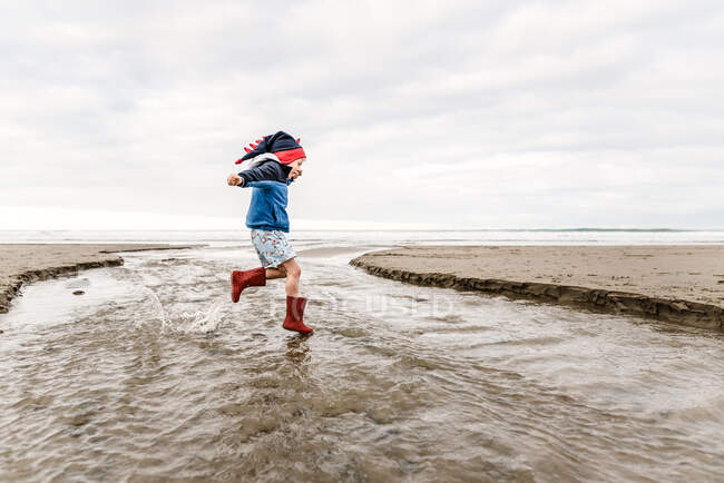 Маленький ребенок в вязаной шляпе динозавра, прыгающий на пляже — стоковое фото