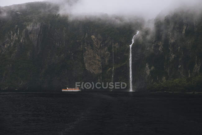 Ferry turístico acercándose cascada en Milford Sound durante el día brumoso, Nueva Zelanda - foto de stock