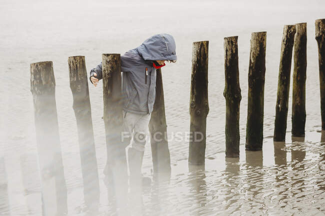 Visão de comprimento total do menino apoiando-se em pilhas em uma praia fria de inverno — Fotografia de Stock
