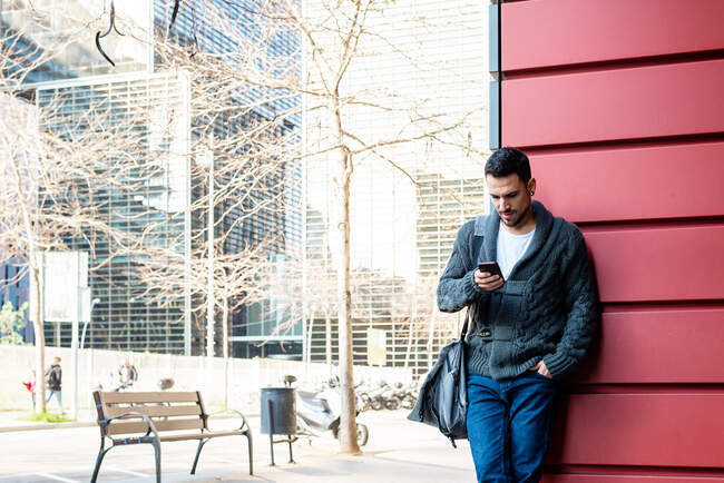 Front view of a bearded man using phone leaning on office building wall — Stock Photo