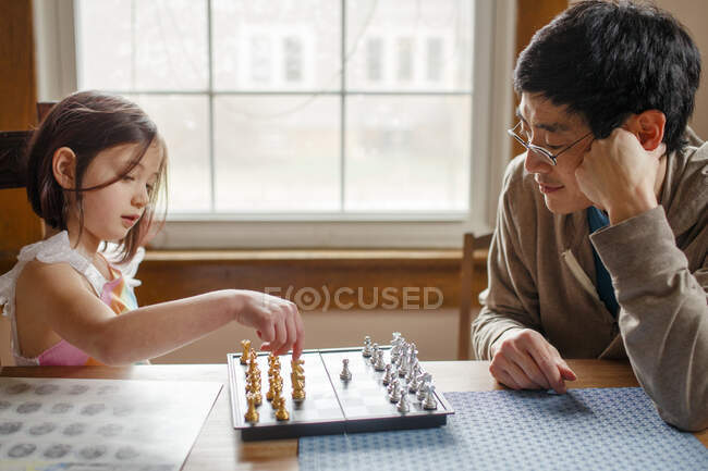 Маленький ребенок играет в шахматы с улыбающимся отцом в свете окон — стоковое фото