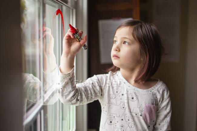 Une petite fille sérieuse dessine sur une fenêtre avec un marqueur rouge vif — Photo de stock