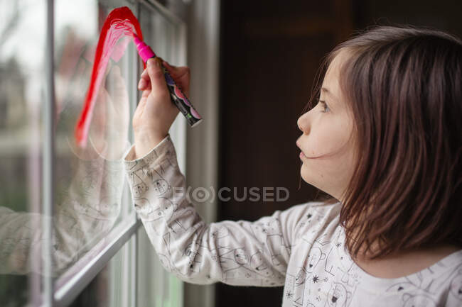 Un bambino piccolo e serio disegna un arcobaleno su una finestra con un pennarello rosso — Foto stock