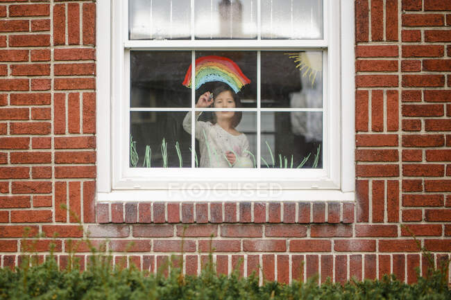 Вид снаружи маленького ребенка, стоящего в окне и рисующего радугу — стоковое фото