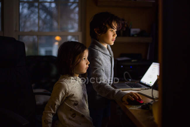 Dos niños se paran en una habitación oscura con las caras iluminadas por una pantalla de ordenador - foto de stock