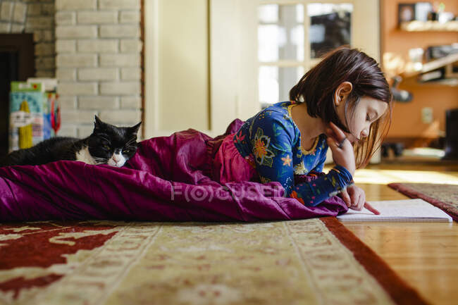 Un enfant se recroqueville sur le sol dans un sac de couchage avec un chat faisant des travaux scolaires — Photo de stock