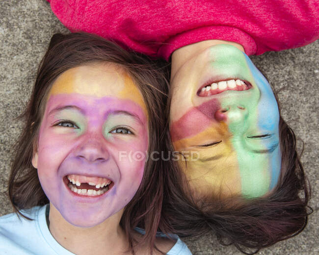 Two children lay down cheek-to-cheek with colorful face paint laughing — Stock Photo