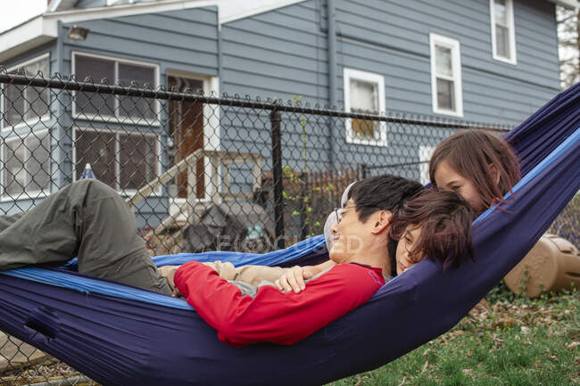 Due bambini felici abbracciano il loro padre su un'amaca nel loro cortile — Foto stock