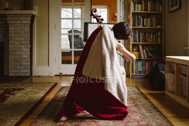 Vue arrière d'un enfant enveloppé dans une couverture rouge pratiquant le violoncelle à l'intérieur — Photo de stock