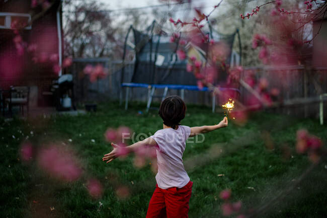 Мальчик стоит под цветущим деревом в сумерках и играет с бенгалькой. — стоковое фото