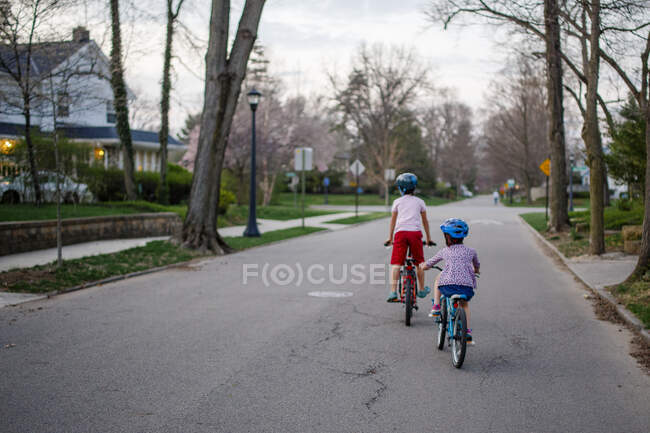 Vista trasera de dos niños en bicicleta juntos por el vecindario al atardecer - foto de stock