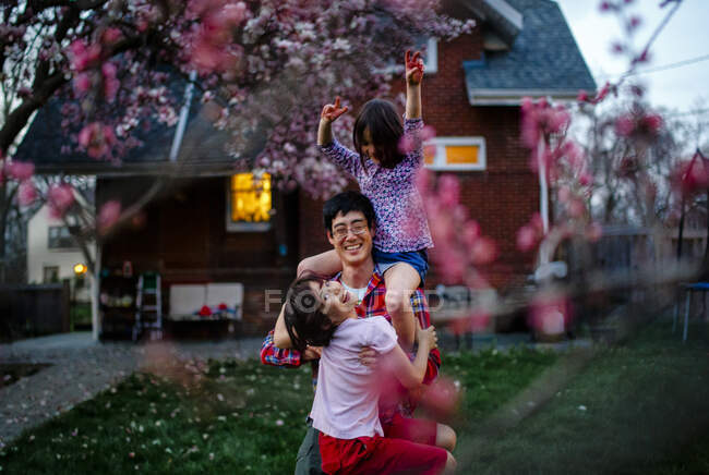 A laughing father carries his two laughing children in yard at dusk — Stock Photo
