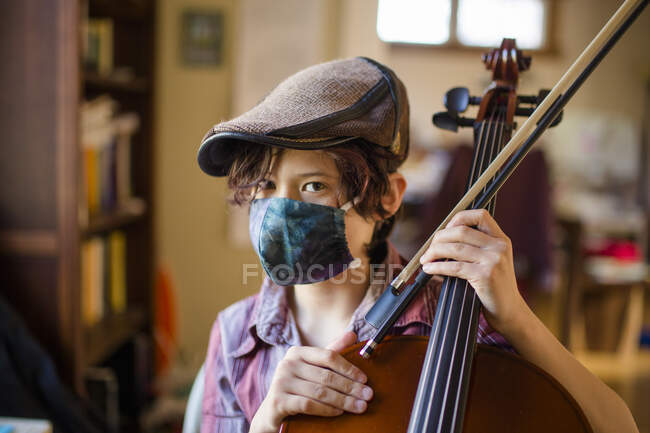 Um menino sério com olhar direto com uma máscara e um boné de lã segura violoncelo — Fotografia de Stock
