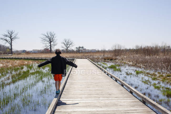 A boy walks on the edge of a wooden bridge across wetlands in Spring — Stock Photo