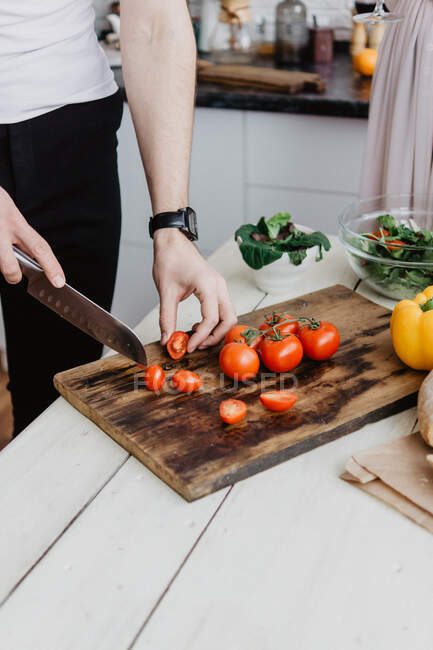 As mãos de homens cortam tomates vermelhos com uma faca em uma tábua de corte — Fotografia de Stock