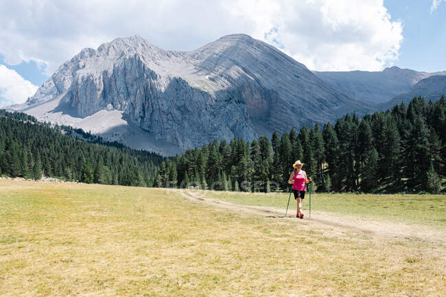 Mujer excursionista con sombrero de paja, pantalones cortos y mochila en el camino a través de una llanura caminando con montañas increíbles en el fondo mientras disfruta del entorno natural alrededor. Fotografía horizontal. - foto de stock