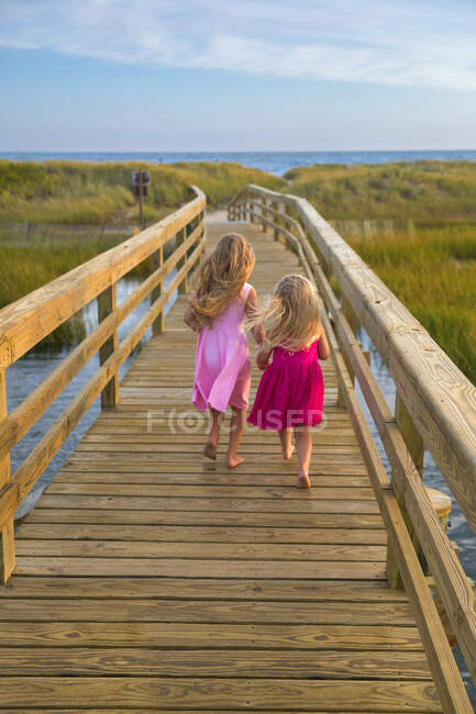 Pequenas meninas por trás correndo na ponte para a praia em vestidos cor de rosa — Fotografia de Stock