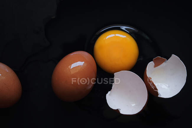 Broken egg with whole brown eggs on a black background, top view — Stock Photo