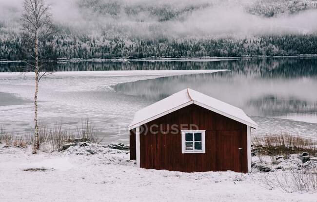 Cabaña roja tradicional con vistas a un lago cubierto de nieve y bosque - foto de stock