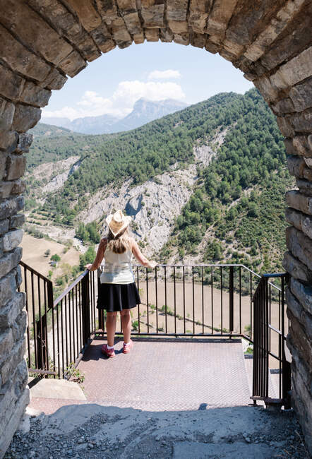 One sexy blonde woman wearing a straw hat, yellow shirt and black skirt close to an iron bar fence looking the landscape of a mountain. The scene is through an ancient stone arch. Vertical ph — Stock Photo