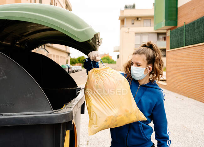 Girl throwing the a yellow trash bag into a green recycling container open in the street. The teenager is wearing face mask and gloves to protect herself from infections, virus, bacterias. Horizontal — Stock Photo