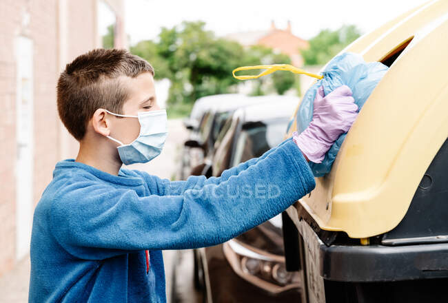 Хлопчик кидає синій мішок для сміття в жовтий контейнер переробки на вулиці. Дитина в масці і рукавичках штовхає сміттєвий мішок, тому що він занадто великий, щоб увійти в смітник. горизонт — стокове фото