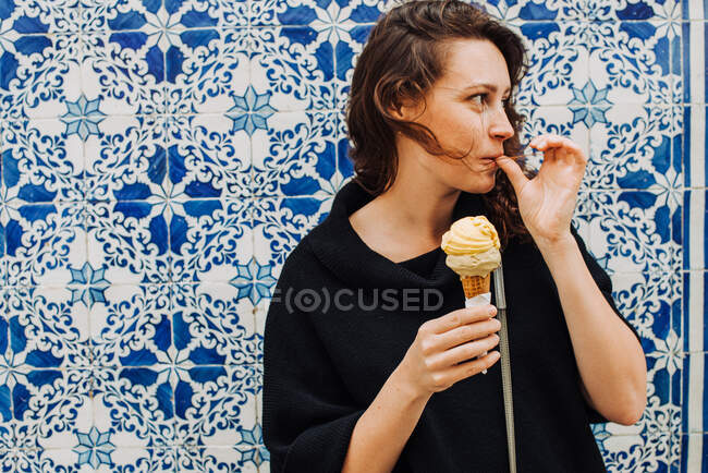 Тысячелетняя женщина облизывает палец, поедая мороженое у плитки на стене — стоковое фото