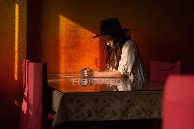 Porträt einer jungen nachdenklichen kaukasischen Frau mit schwarzem Hut, die allein am Tisch im Café vor orangefarbener Wand sitzt — Stockfoto
