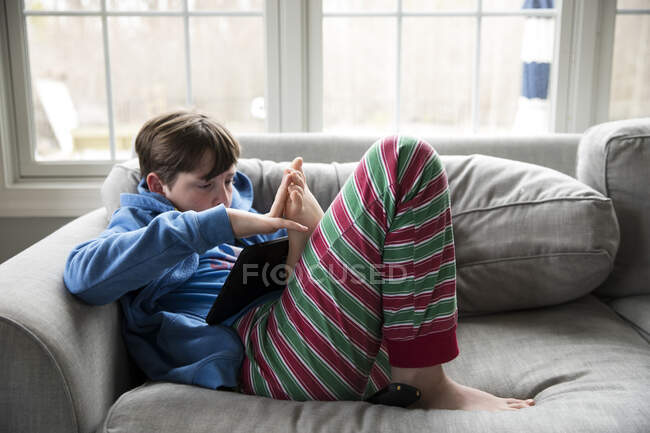 Adolescente ragazzo malato con influenza indossa pigiama a strisce, guardando Ipad sul divano — Foto stock
