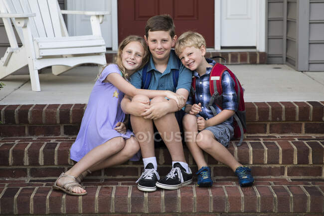Tres hermanos sonrientes se sientan en los escalones del porche delantero de ladrillo - foto de stock