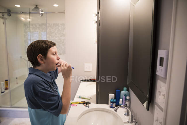 Adolescente menino olha no espelho enquanto escova seus dentes no banheiro moderno — Fotografia de Stock