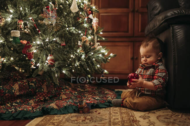 Хлопчик Тоддлер сидить під ялинкою, тримаючи прикрасу — стокове фото