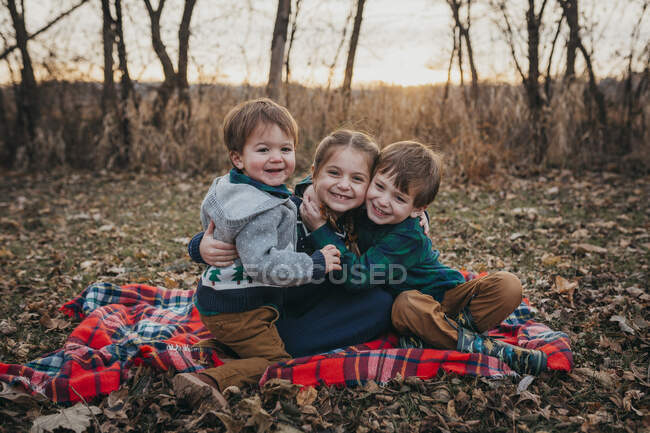 Три брати і сестри сидять на ковдрі, обіймаючись посміхаючись на камеру — стокове фото