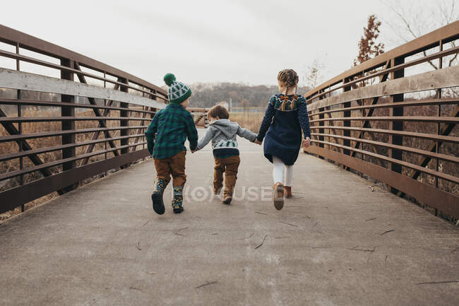 Трое братьев и сестер держатся за руки, бегая по мосту от камеры — стоковое фото